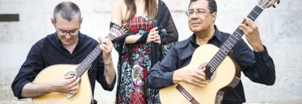 Two older men dressed in black play guitar while a woman in a red dress stands holding her hands behind them.