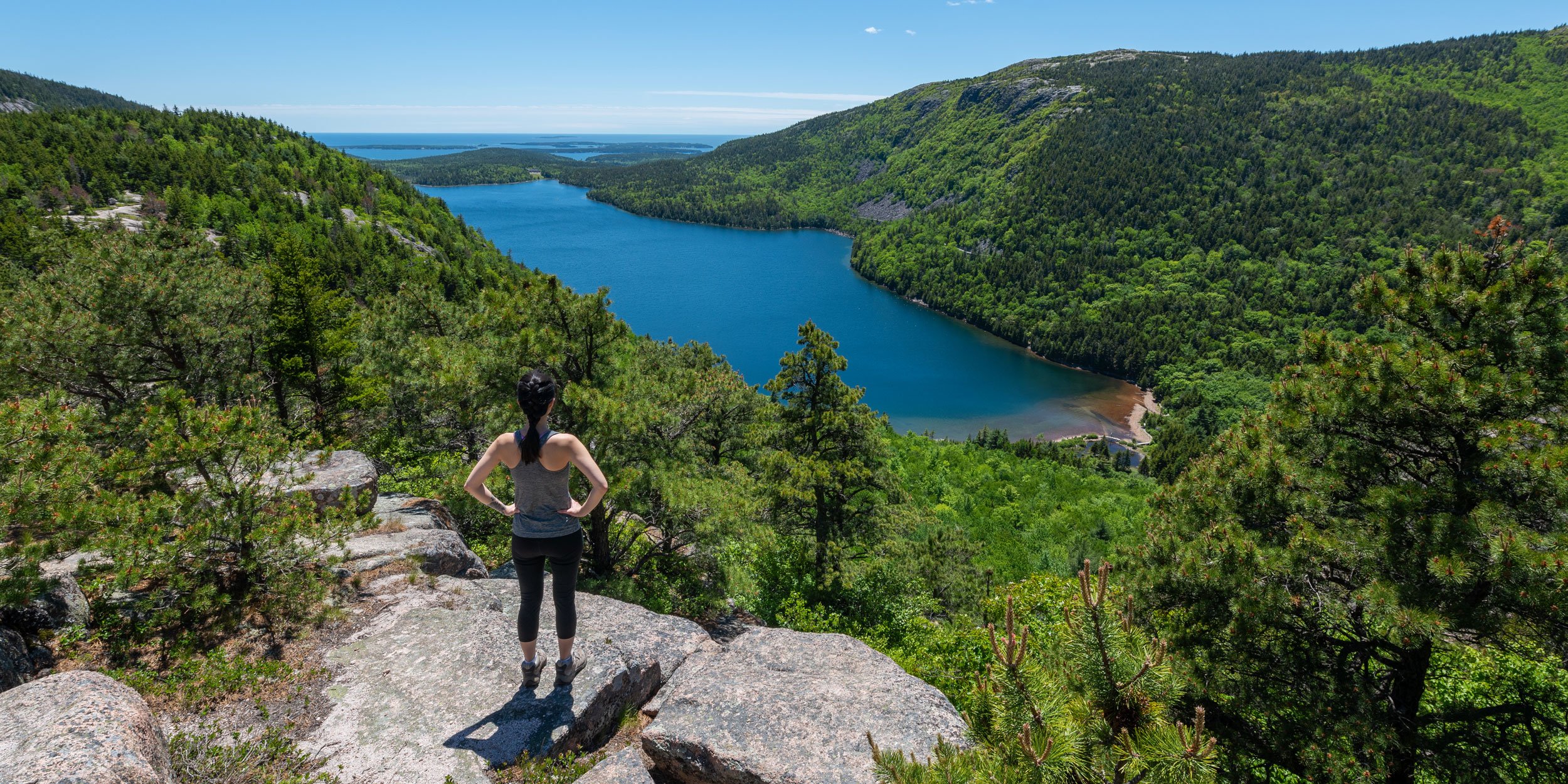 Hiker standing on a rock overlooking water and lush mountains at Acadia National Park, Maine